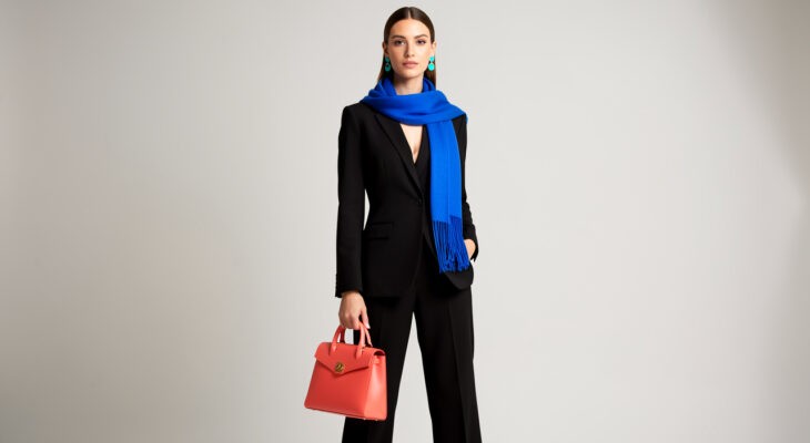 Femme élégante vêtue d'une tenue noire sobre, rehaussée par un sac corail, des boucles d'oreilles turquoise et une écharpe bleu cobalt.